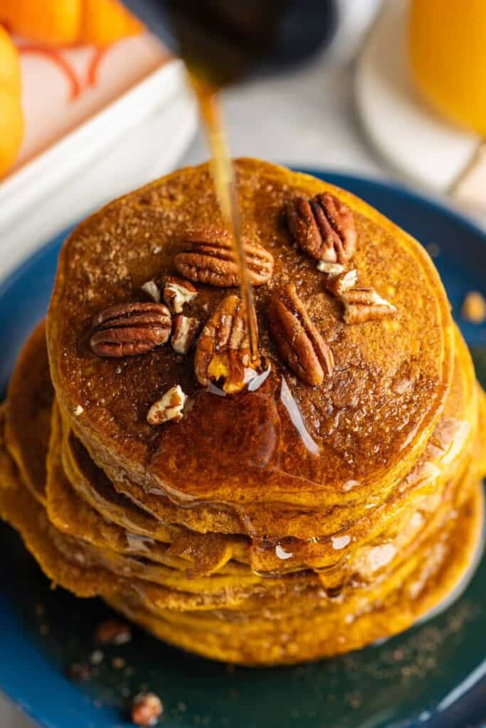Maple syrup pouring over gluten free pumpkin pancakes stacked up on a plate with pecans on top.
